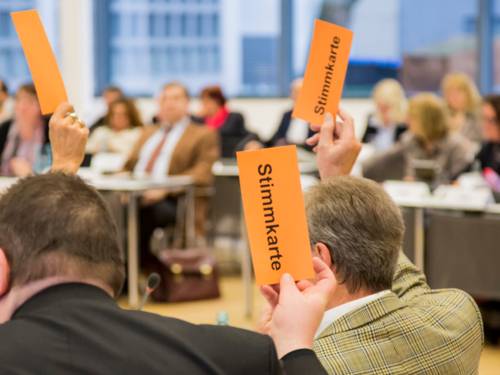 Mitglieder der Regionsversammlung heben orangefarbene Pappkarten mit dem schwarzen Text "Stimmkarte" in die Luft.