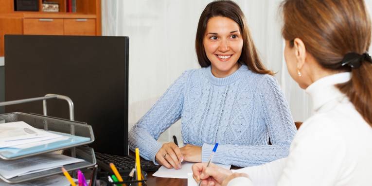 Beratungssituation in einem Büro, bei der sich zwei Frauen mit Stift und Zettel an einem Schreibtisch besprechen.